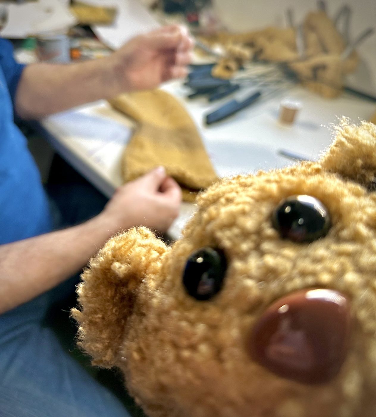 A handmade puppet character peering into the camera on a busy workshop table